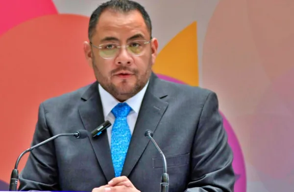 Hombre con barba, lentes y corbata azul habla frente a dos micrófonos durante una conferencia de prensa.